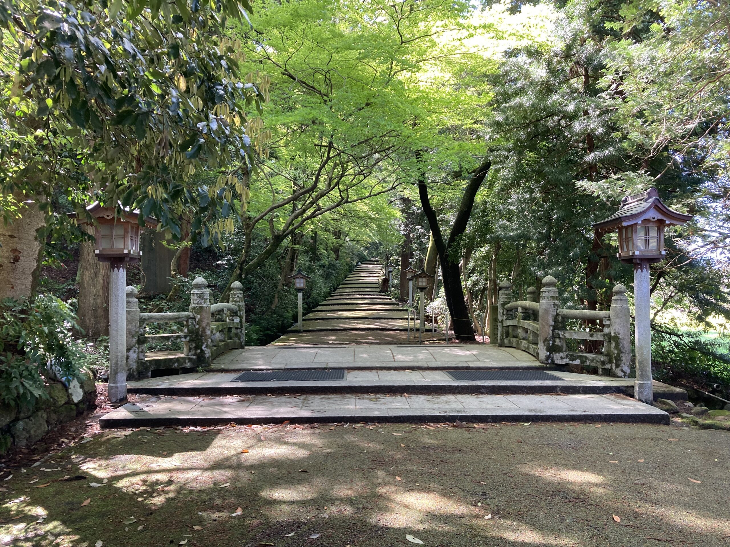 Stone footbridge with traditional lanterns along a mossy stone path surrounded by lush green trees and shadows.